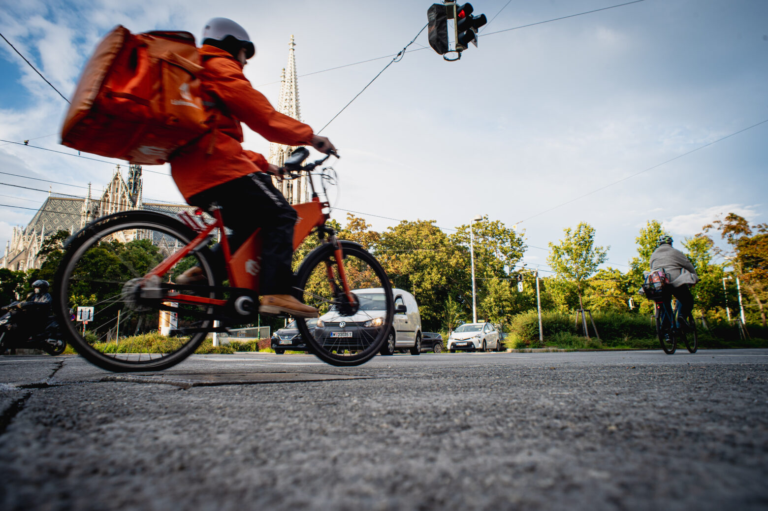 Ein Essenslieferant fährt mit dem Fahrrad über eine Kreuzung in Wien. Im Hintergrund ist die Votiv-Kirche zu sehen. Das Fahrrad, die Jacke und der Rucksack des Lieferanten sind im bekannten Orange von Lieferando.