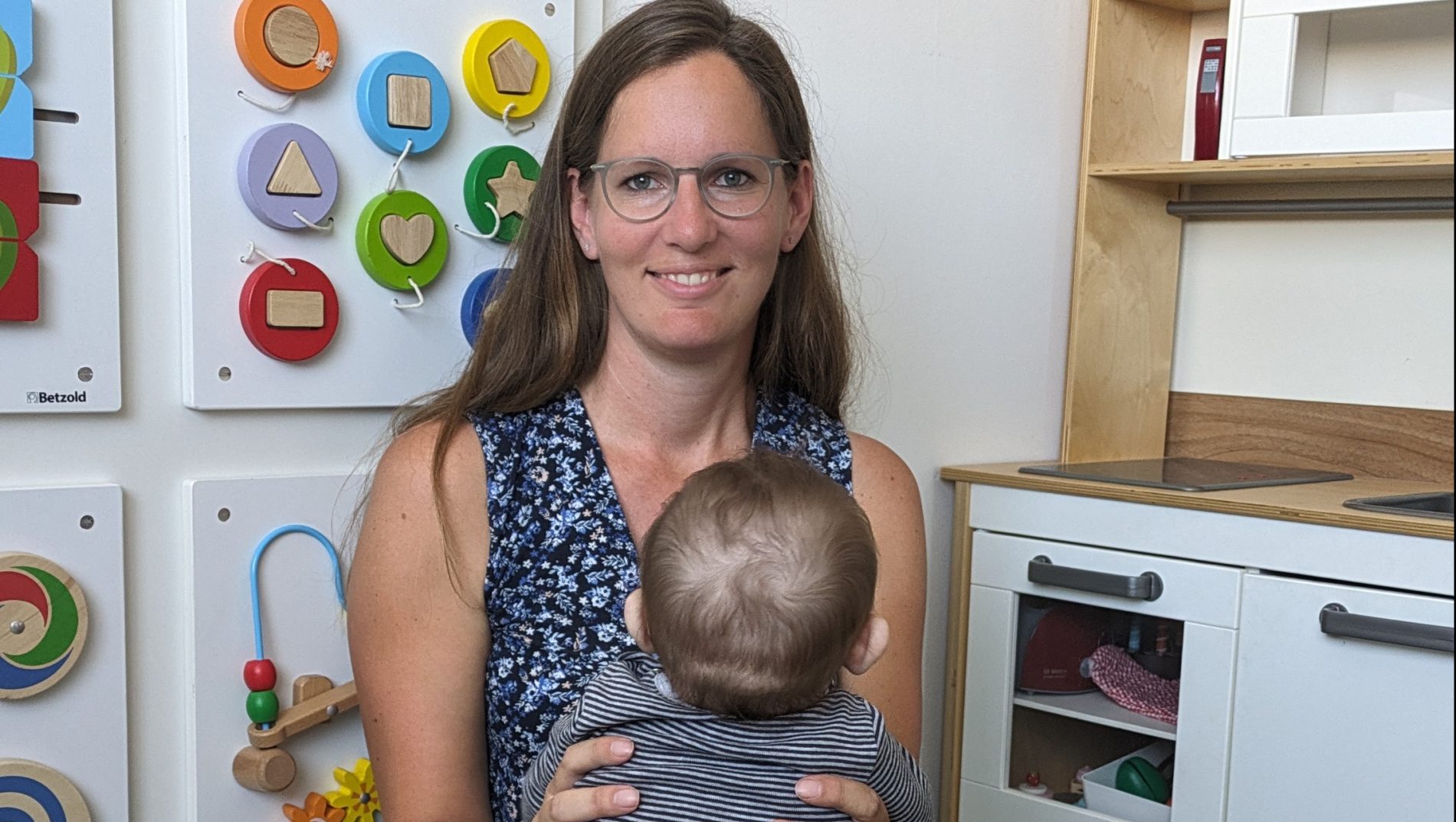 Krisenpflegemutter Raab mit einem von hinten zu sehenden Pflegekind im Arm in einem Kinder-Spielzimmer.